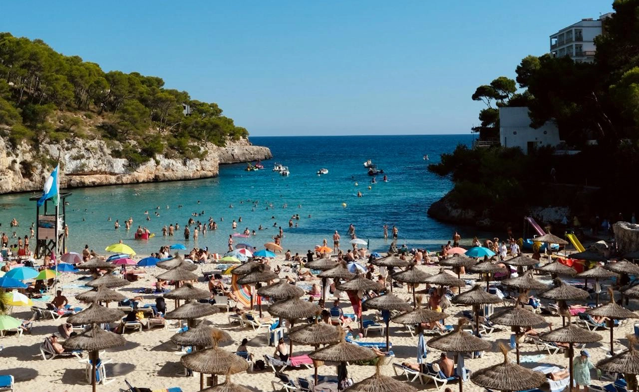 A crowded beach in Spain with turquoise waters, surrounded by cliffs and lush greenery, perfect for relaxation and golf enthusiasts exploring Golf Spain.