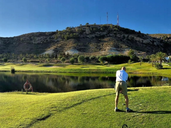 Picturesque golf course at one of Spain's best golf resorts, featuring a lush green fairway, serene lake, and dramatic mountain backdrop. A golfer prepares to tee off, showcasing the world-class golfing experience available at top Spanish golf destinations.