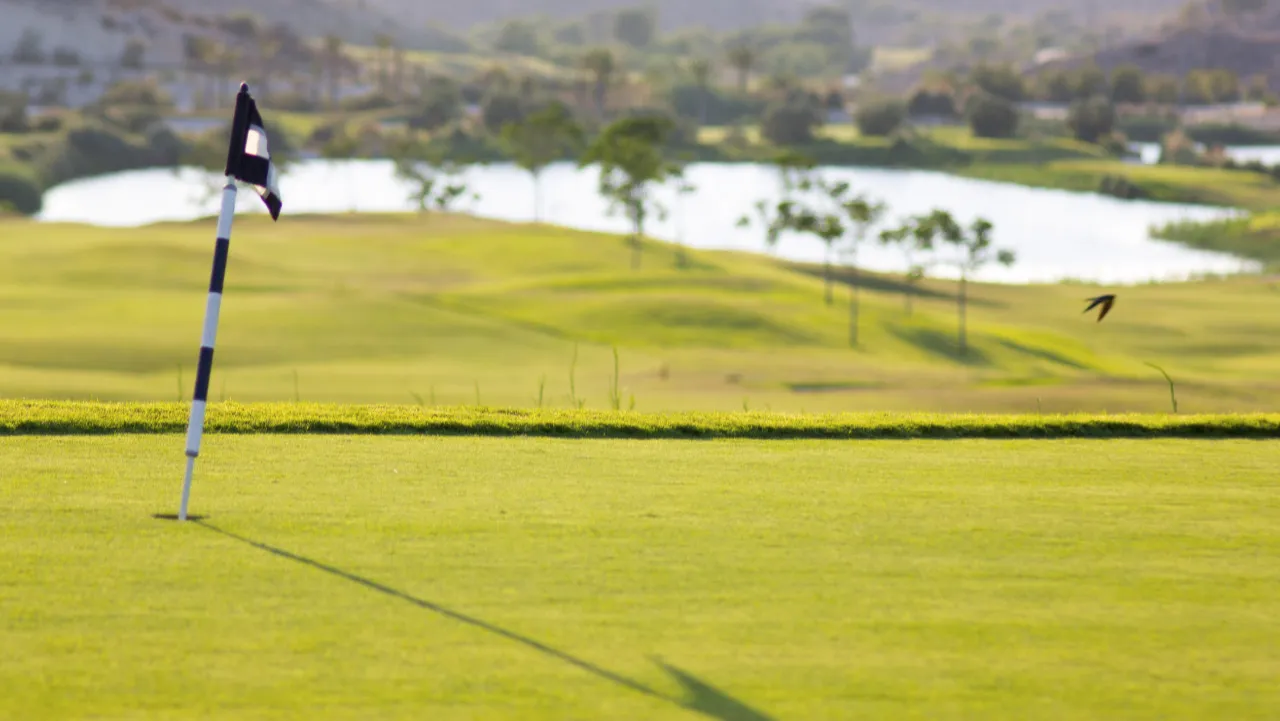 Scenic golf course in Murcia, Spain, with lush green fairway, flag on putting green, rolling hills, and water hazard in background