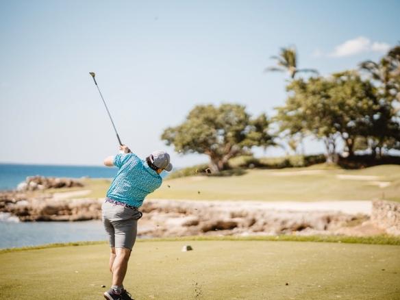 A golfer in a turquoise patterned shirt takes a swing on a scenic coastal golf course with palm trees and rocky shoreline, reminiscent of luxury golf breaks in Spain.