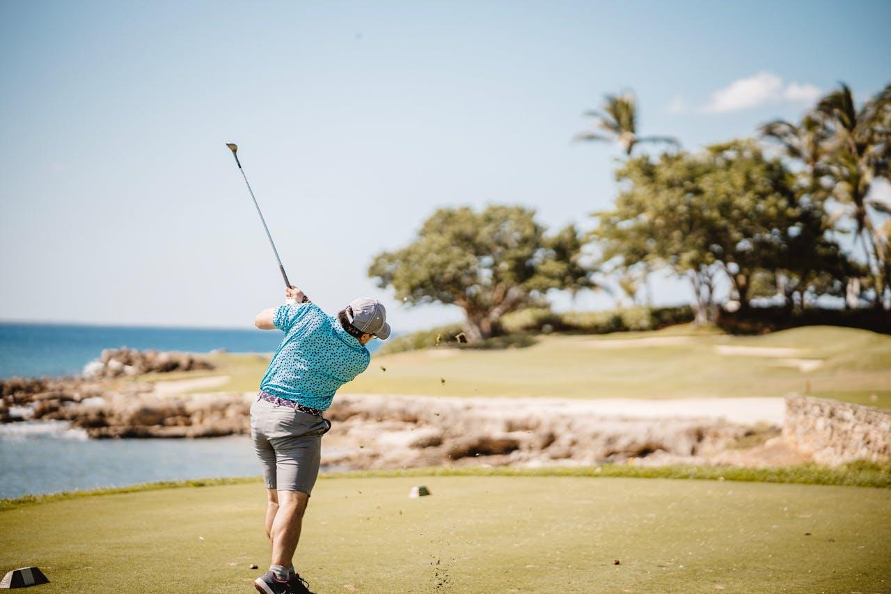 A golfer in a turquoise patterned shirt takes a swing on a scenic coastal golf course with palm trees and rocky shoreline, reminiscent of luxury golf breaks in Spain.